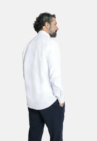 Camisa blanca de manga larga con cuello puntiagudo, que presenta una textura suave y un dobladillo curvado, combinada con pantalones azul oscuro.