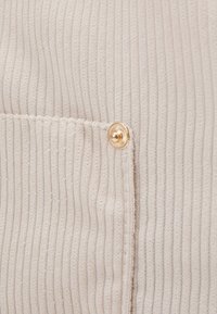 Tissu beige côtelé à la texture douce, avec un bouton rond doré près d'une poche cousue.