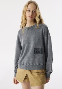 Felpa grigia in cotone con collo rotondo, spalle scese e tasca frontale con testo nero in rilievo. Abbinata a pantaloni corti khaki.