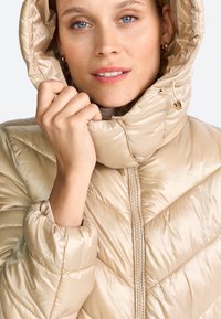 Beige opblaasjack met hoge kraag en capuchon, voorzien van een rits aan de voorkant, elastische manchetten en een gladde, glanzende textuur.