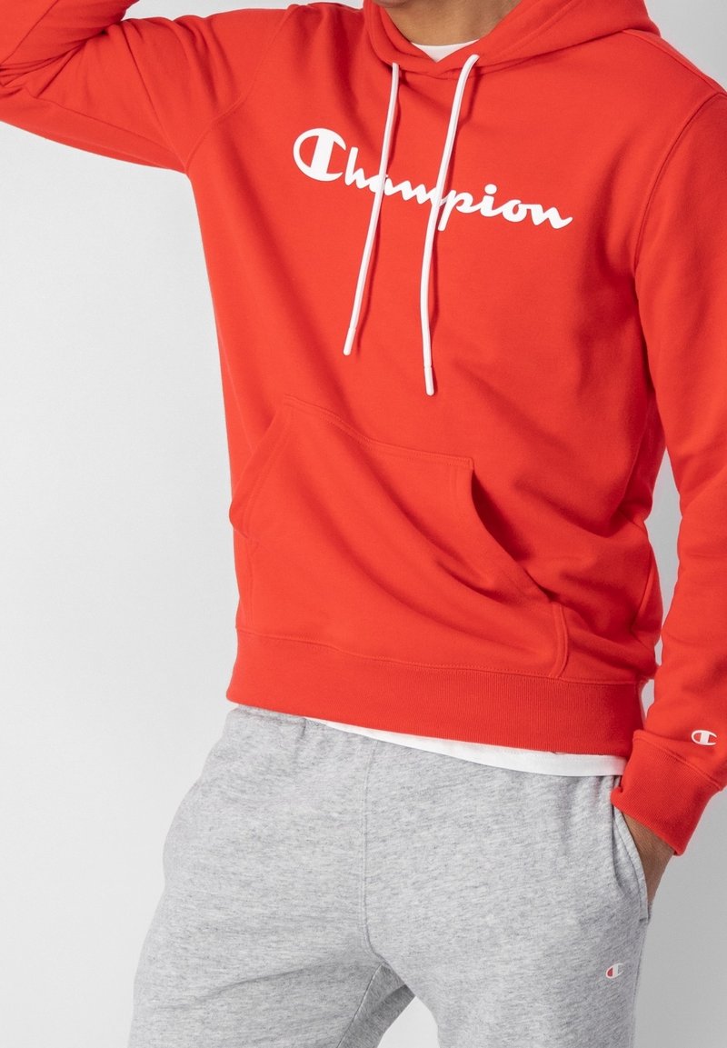 Czerwona bluza Champion z białym logo, sznurkami i kieszenią z przodu. W zestawie z szarymi dresami z logo na nogawce. Casualowy strój sportowy.