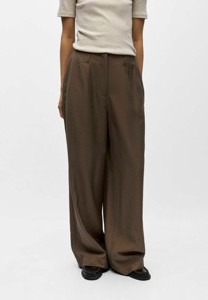 Personne portant un pantalon large marron taille haute et un haut beige à manches courtes, debout avec les mains détendues le long du corps.