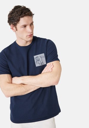 Uomo con capelli castani ricci che indossa una maglietta blu navy con un disegno astratto di un cavallo sul petto, braccia incrociate, guarda di lato.