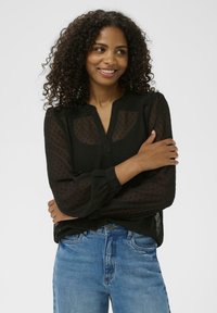 Zwarte doorzichtige blouse met een subtiel stippenpatroon, lange mouwen en een knoopsluiting aan de voorkant, gecombineerd met blauwe denim jeans.