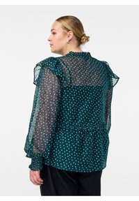 Türkise Bluse mit transparenter gepunkteter Überlagerung, Rüschenschultern und gestuften Saum. Verfügt über einen subtilen Glanz und ein zartes gepunktetes Muster.