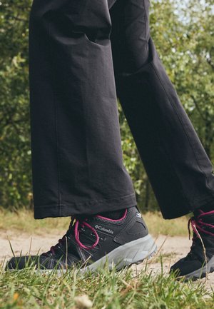 Personne portant des chaussures de randonnée Columbia noires avec des lacets roses et un pantalon noir marchant sur un chemin extérieur herbeux.