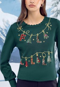 Zeleni pleteni pulover s čipkastim izrezom, okrašen s prazničnim vezenim napisom "FA LA LA" in dekoracijo iz bleščic, vključno z drevesci in zvezdami.