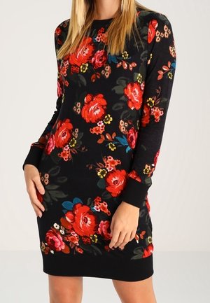 Woman wearing a black long-sleeve dress with a red, pink, and yellow floral pattern, standing with hands near hips.