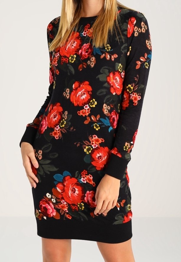 Femme portant une robe noire à manches longues avec un motif floral rouge, rose et jaune, debout les mains près des hanches.