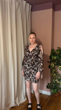 Νεαρή γυναίκα με μίνι φόρεμα μακρυμάνικο με λουλουδάτο σχέδιο και ανοίγματα στους ώμους, στέκεται μέσα σε εσωτερικό χώρο δίπλα σε μπεζ κουρτίνες και μια γλάστρα με φυτό.