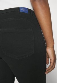 Los pantalones de mezclilla negros presentan un bolsillo trasero con forma puntiaguda. Una etiqueta azul en la cintura muestra "ONLY CARAMKOMA Denim." Textura suave en general.