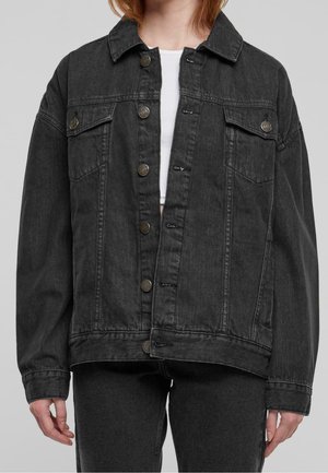 Henkilö, jolla on musta farkkutakki metallisilla napeilla valkoisen lyhyen topin päällä, yhdistettynä mustiin farkkuihin, seisoo harmaan taustan edessä.