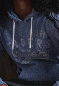 Sudadera azul de tejido suave, con un bolsillo tipo canguro, cordones blancos y una impresión gráfica frontal con "SuperDry" y "TIGERS."