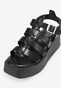 Sandalia de plataforma de cuero negro con tiras trenzadas, punta cuadrada, suela gruesa y hebilla ajustable. Textura lisa y diseño minimalista.