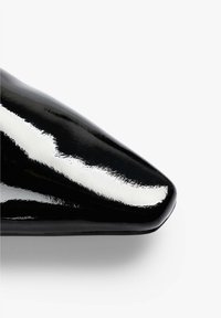 Zapato de punta afilada en charol negro con un acabado brillante. Presenta un diseño elegante y una textura suave, que enfatiza su silueta definida.