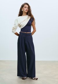 Pantaloni larghi blu navy con pieghe, abbinati a una maglietta aderente blu navy e a un maglione crema gettato su una spalla. Scarpe marroni con fantasia.