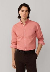 Hellkorallenfarbenes Hemd mit langen Ärmeln, Kragen und Brustlogo, kombiniert mit dunkelbrauner Hose und schwarzem Gürtel.