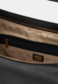 Intérieur d'un sac à main en cuir noir avec une doublure beige ornée du logo "GUESS" répété, une fermeture éclair dorée et une poche intérieure zippée.