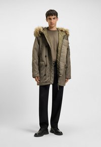 Olijfgroene parka met een nepbont gevoerd capuchon, dubbelknopen, zijzakken en een relaxte pasvorm, gecombineerd met zwarte broeken en schoenen.