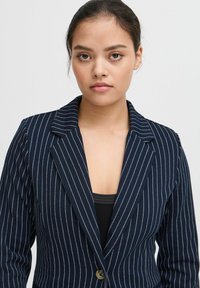 Blazer bleu marine à rayures fines avec un col cranté, fermeture à un bouton et une texture lisse, porté sur un haut noir.