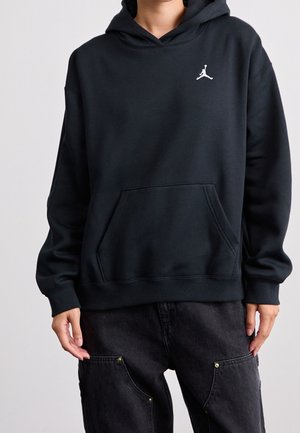 Personne portant un sweat à capuche noir avec un logo Jumpman blanc sur la poitrine et un pantalon cargo noir sur un fond uni.