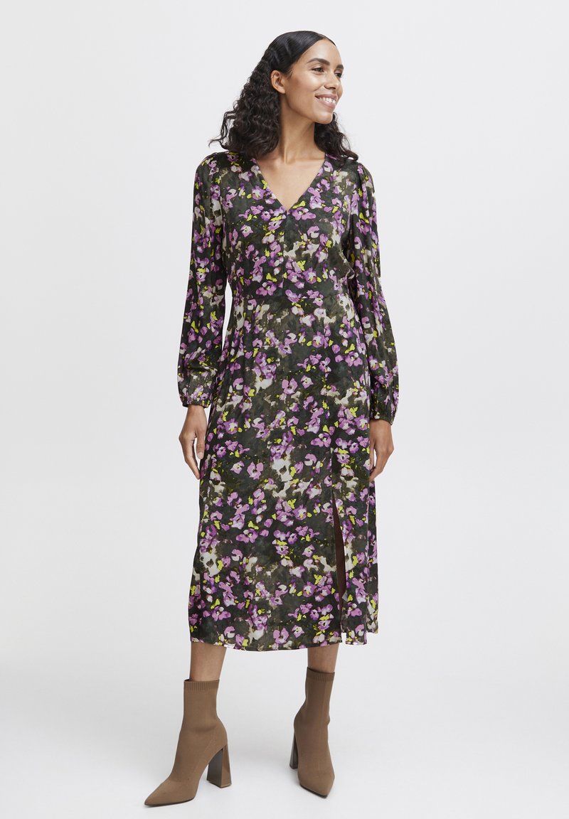 Robe midi à motifs floraux avec un motif vert foncé et violet, décolleté en V profond, manches longues bouffantes et une fente sur le côté. Associée à des bottes à talons brunes.