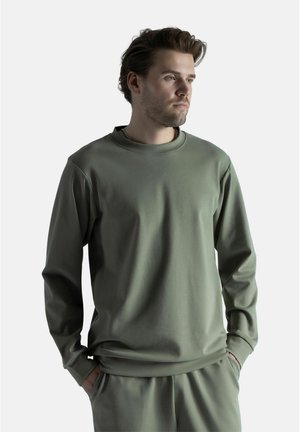 Mies, jolla on lyhyet ruskeat hiukset, pukeutunut yksiväriseen oliivinvihreään pitkähihainen paitaan ja siihen sopiviin housuihin, kädet taskuissa, katsoo oikealle.