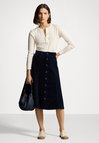 Mørkeblå corduroy midi-nederdel med frontknaplukning, parret med en lysegrå langærmet top og beige sneakers, accessoriseret med en mørk taske.