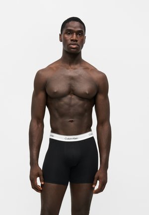 Мускулест тъмнокож мъж стои, облечен в черни боксерки Calvin Klein с бяла лента около талията върху едноцветен светъл фон.