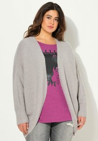 Světle šedý pletený kardigán s volnějším střihem a žebrovanými manžetami, vrstvený přes fialové tričko s potiskem, které má černý design, spárovaný s potrhanými džíny.