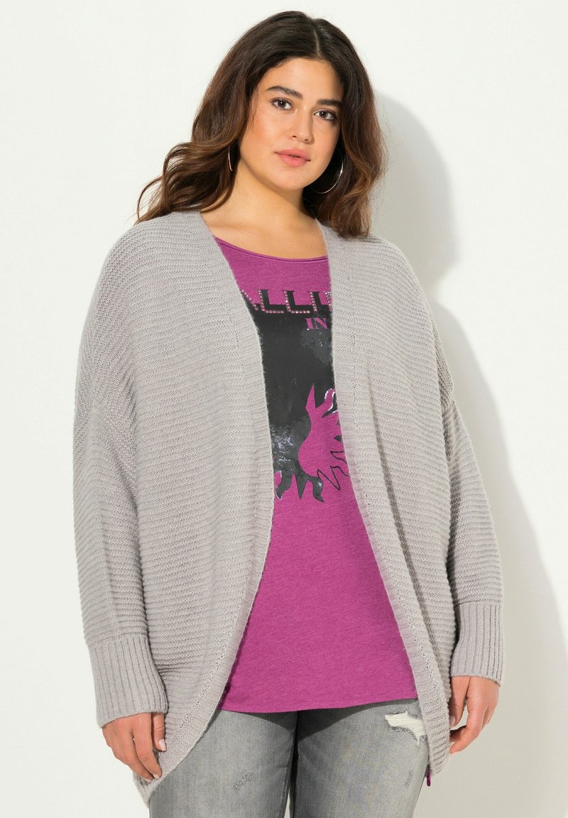 Světle šedý pletený kardigán s volnějším střihem a žebrovanými manžetami, vrstvený přes fialové tričko s potiskem, které má černý design, spárovaný s potrhanými džíny.