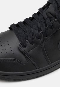 Gros plan d'une basket en cuir noir avec de gros lacets noirs et des perforations sur la zone des orteils pour la respirabilité.