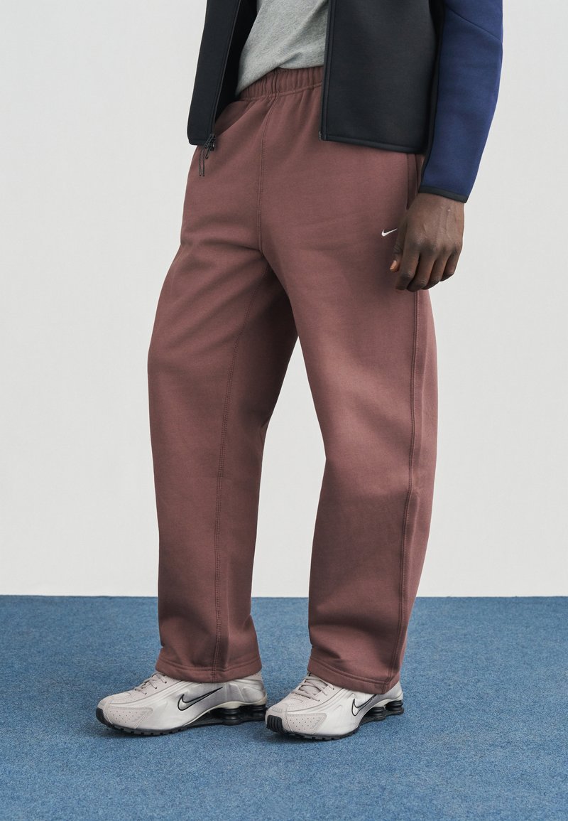 Nike laisvalaikio kelnės švelnios bordo spalvos, su elastine juosmens juosta, šoninėmis kišenėmis ir subtiliu logotipu. Derinamos su pilkomis Nike sportinėmis avalynėmis.