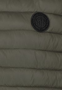 Nahaufnahme von olivgrünem, gestepptem Jackenstoff mit horizontalen Nähten und einem runden, schwarzen Markenpatch, der aufgenäht ist.