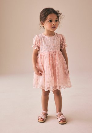 Young girl with curly hair wearing a pink floral lace dress and matching sandals, standing against a beige background.