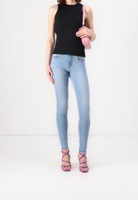 Černý žebrovaný top bez rukávů, světle modré úzké džíny a fialové sandály na podpatku se stříhacími pásky. Modelka drží malou růžovou kabelku. Jednoduché pozadí.