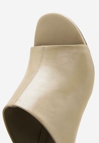 Chaussures à talons compensés beiges avec orteils ouverts, design incurvé, texture de cuir lisse et détails de coutures visibles sur le côté.