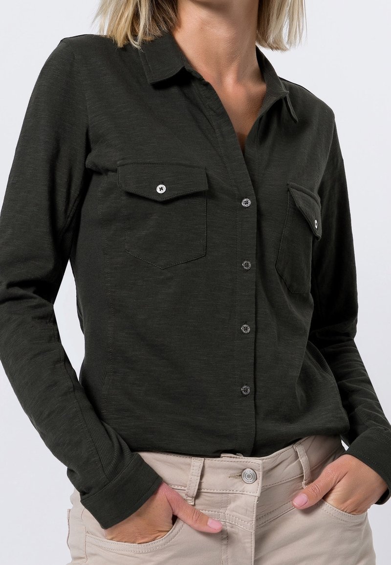 Femme portant une chemise boutonnée verte foncée avec des poches poitrine et un pantalon beige, les mains dans les poches, sur un fond uni.