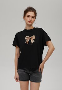 Camiseta de algodón negra con mangas cortas que presenta un gráfico de un lazo con estampado de leopardo en la parte frontal. Combinada con pantalones cortos de mezclilla grises. Textura suave.
