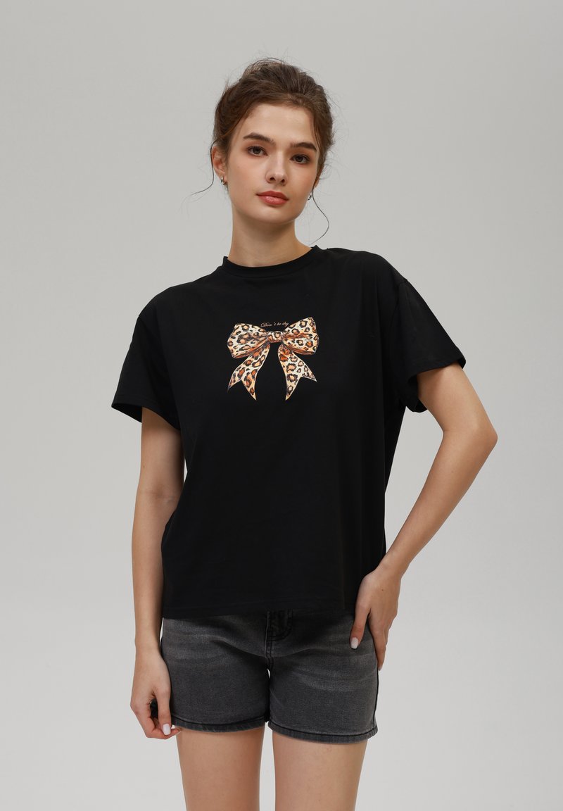 Camiseta de algodón negra con mangas cortas que presenta un gráfico de un lazo con estampado de leopardo en la parte frontal. Combinada con pantalones cortos de mezclilla grises. Textura suave.