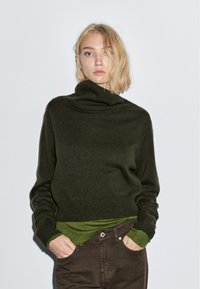 Dunkelgrüner, grob gestrickter Rollkragenpullover mit einem verkürzten Design. Über einem hellgrünen Langarmshirt getragen, kombiniert mit braunen Hosen.