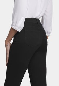 Jeans de mezclilla negra con un diseño ajustado, que cuentan con dos bolsillos traseros, una cintura alta y una textura suave. La modelo lleva una blusa blanca.