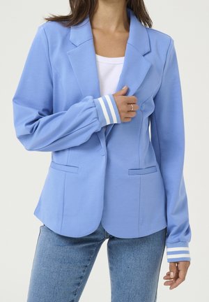 Mujer que lleva un blazer azul claro con puños blancos a rayas sobre una camisa blanca y vaqueros azules, sosteniendo la solapa del blazer con una mano.