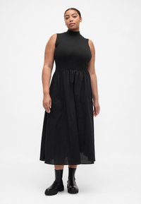 Schwarzes ärmelloses Kleid mit hohem Ausschnitt, eng anliegendem Oberteil und ausgestelltem Rock. Kombiniert mit schwarzen Stiefeletten. Glatter Stoff mit dezentem Schimmer.