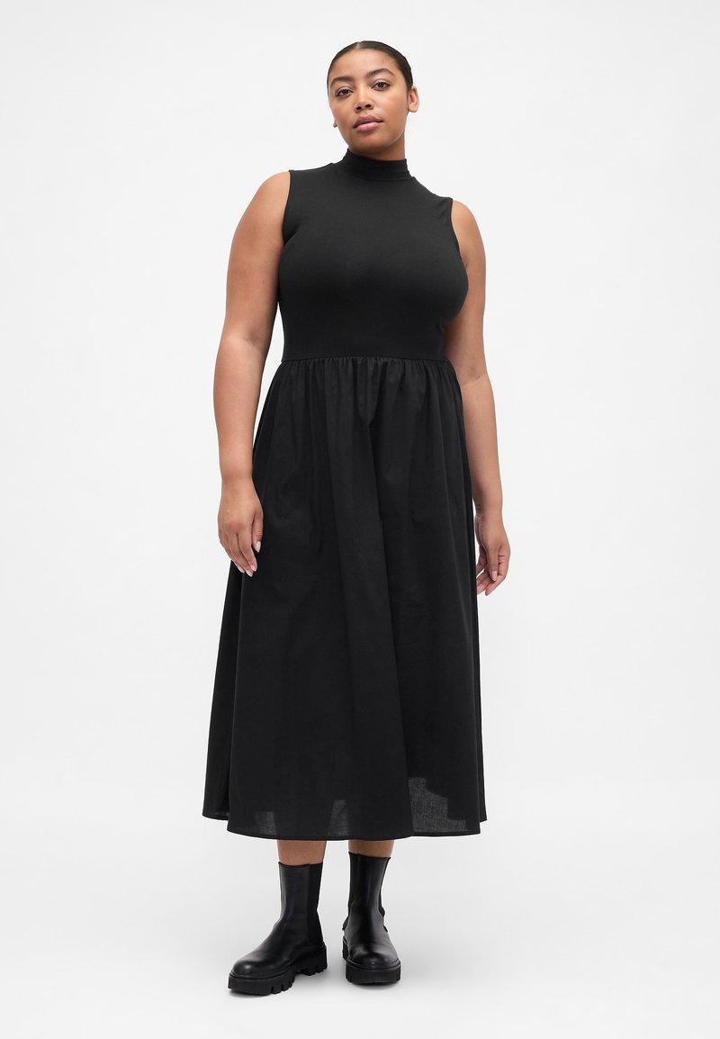 Schwarzes ärmelloses Kleid mit hohem Ausschnitt, eng anliegendem Oberteil und ausgestelltem Rock. Kombiniert mit schwarzen Stiefeletten. Glatter Stoff mit dezentem Schimmer.