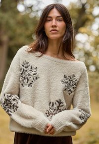 Cremefarbener, flauschiger Pullover mit schwarzen Blumenmustern, weitem Ausschnitt und langen Ärmeln. Weiche Textur, entspannte Passform, leichtes Design.