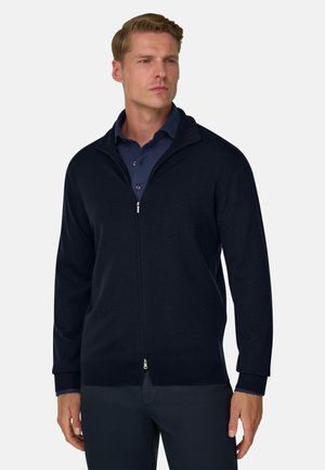 Homme portant un pull zippé bleu marine foncé par-dessus un polo bleu marine, debout avec une expression neutre devant un fond clair uni.