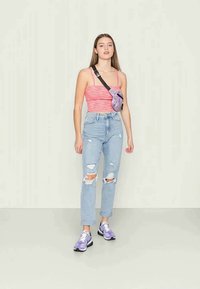 Top sem mangas em cor-de-rosa com vinco, jeans rasgados em azul claro, sapatilhas roxas e pretas, e uma pochete multicolorida usada atravessada pelo corpo.