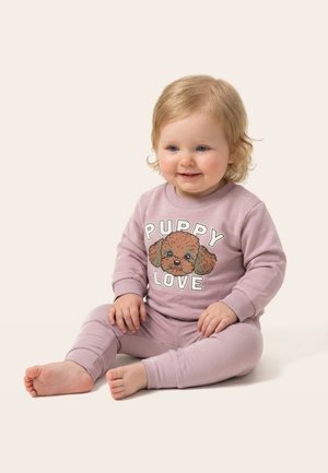 Copil mic cu păr blond care stă pe podea, purtând un costum roz cu fața unui cățeluș și textul „PUPPY LOVE” pe partea din față.
