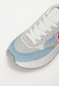 Lichtgrijze sneaker met blauwe en witte accenten, messtof, textuur-overlay en platte veters. Opvallend roze logo aan de zijkant.